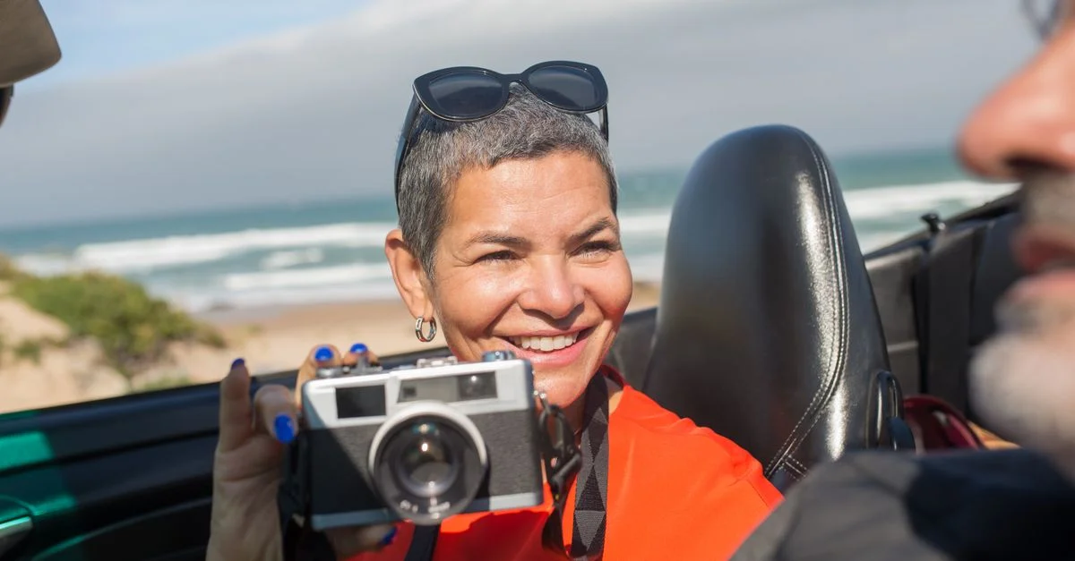 Femme dans un cabriolet souriante, capturant des moments de plage avec un appareil photo vintage.
