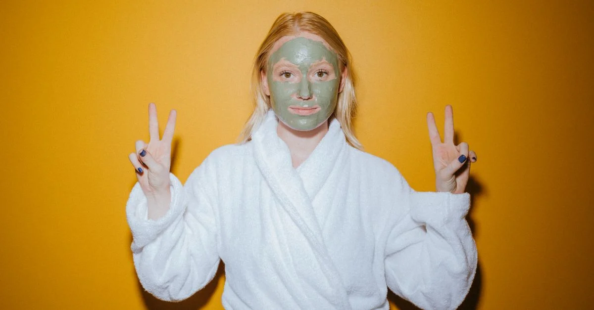 Une jeune femme avec un masque facial pose en peignoir, faisant des signes de paix, devant un fond jaune vibrant.