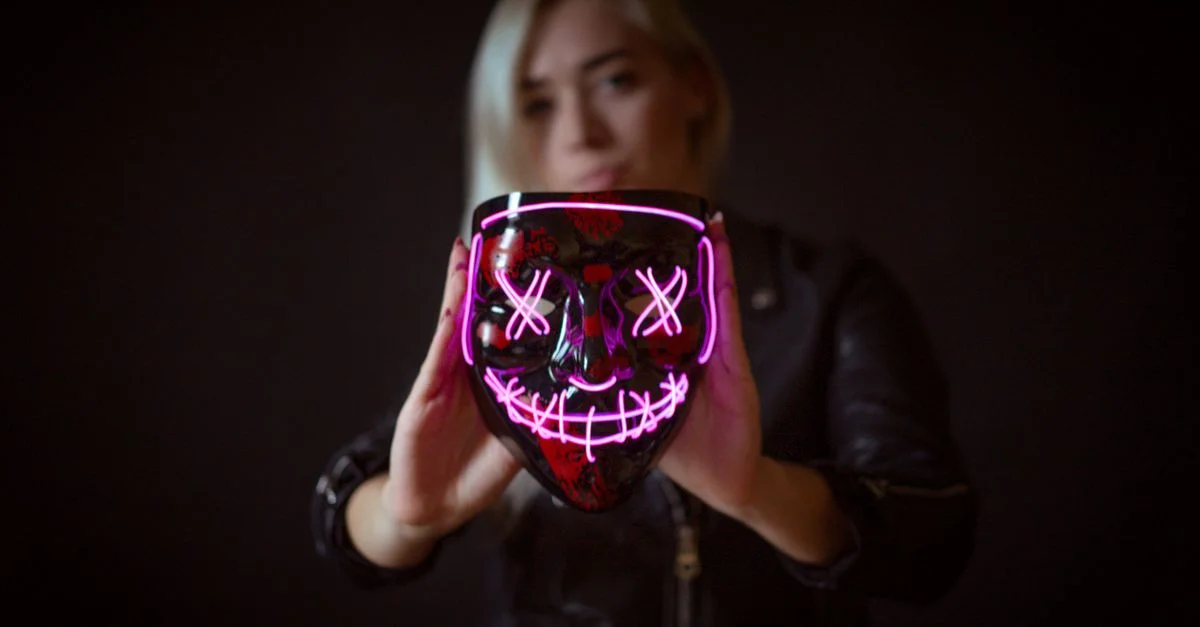 Une femme blonde en veste noire tient un masque LED néon lumineux contre un fond noir.