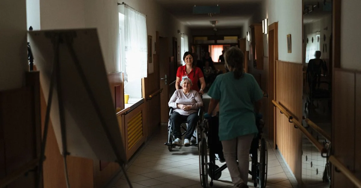 Aide-soignant assistante une femme âgée dans un fauteuil roulant à travers le couloir d'une maison de retraite.