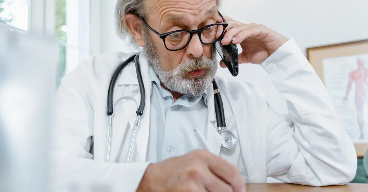 Médecin âgé au bureau au téléphone avec un patient, prenant des notes.