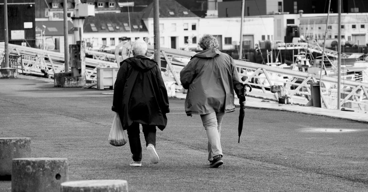 Deux personnes âgées marchent le long du front de mer d'une ville avec des bâtiments en arrière-plan.
