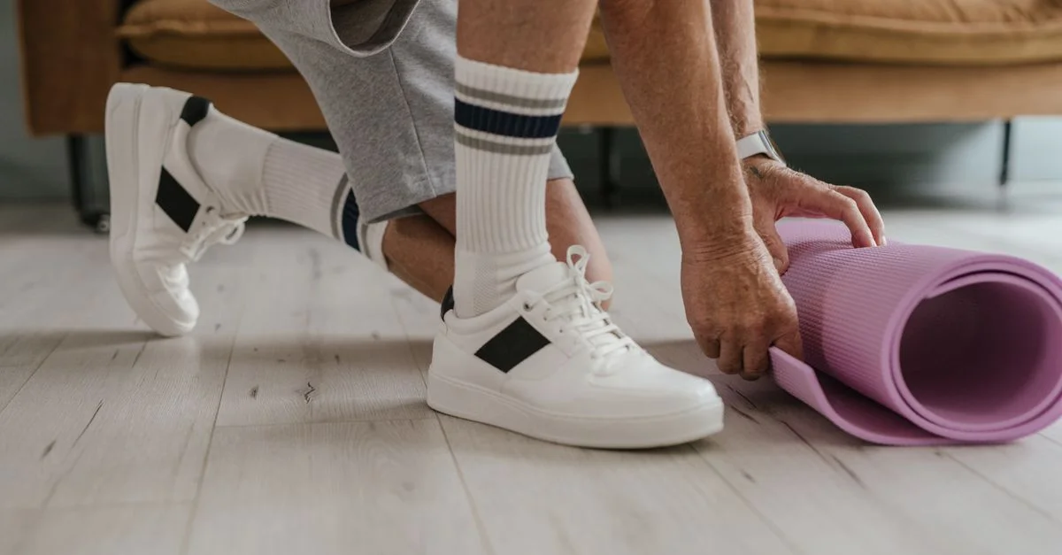 Homme senior en baskets roulant un tapis de yoga violet sur un sol en bois à l'intérieur.
