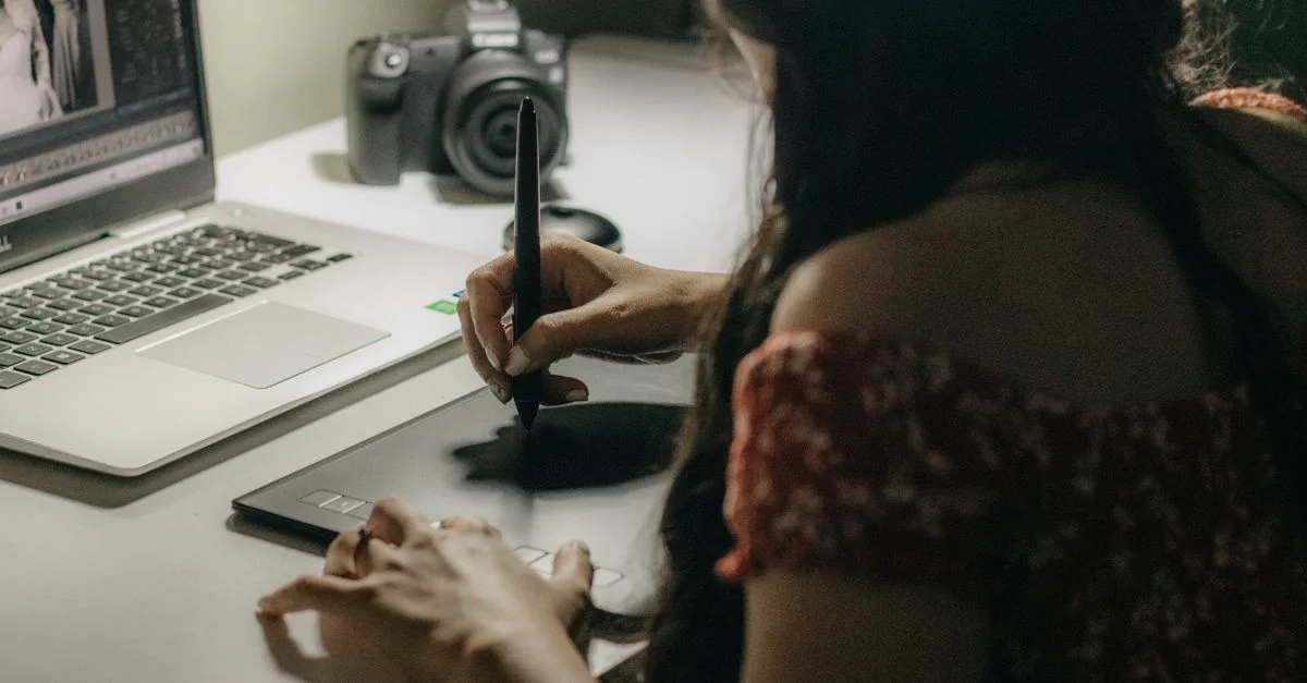 Une femme adulte utilisant une tablette graphique tout en travaillant sur un ordinateur portable dans une pièce faiblement éclairée.