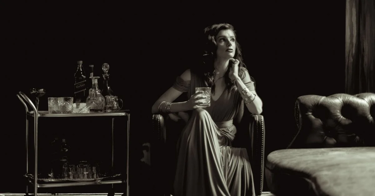 Une femme en robe fluide s'assoit élégamment avec un verre dans un décor vintage en noir et blanc.