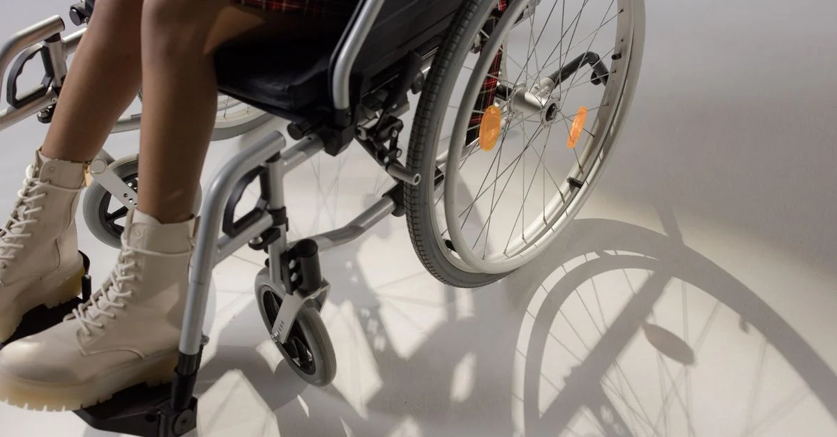 Une jeune femme assise dans un fauteuil roulant, portant des bottes beiges élégantes, projetant une ombre.