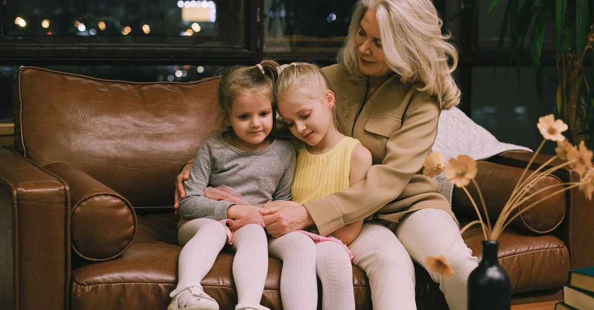 Une grand-mère étreint tendrement ses deux petites-filles sur un canapé en cuir confortable à l'intérieur.