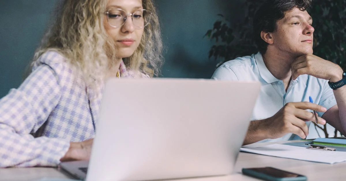 Deux collègues collaborent à un bureau avec des ordinateurs portables et des papiers, suggérant travail d'équipe et productivité.