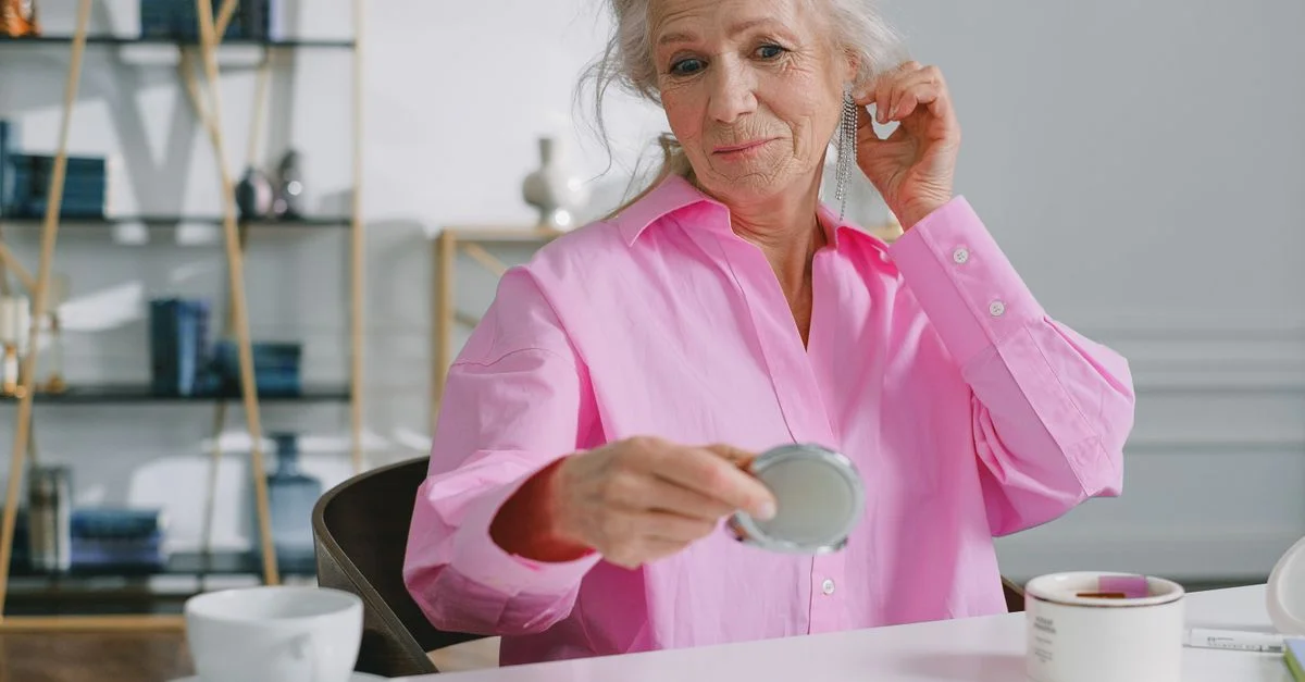 Une femme senior en chemise rose utilisant un miroir de poche pour admirer ses boucles d'oreilles à l'intérieur.
