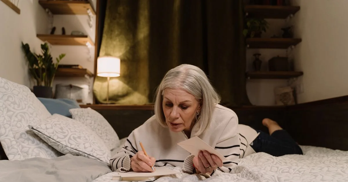 Femme senior allongée sur un lit en train d'écrire une lettre dans une chambre confortable et douillette.