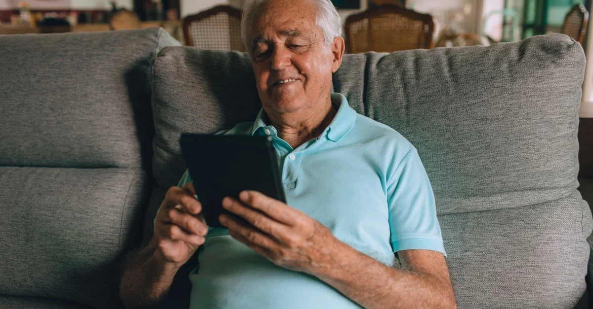 Homme âgé en polo bleu utilisant une tablette sur un sofa, souriant à l'intérieur.