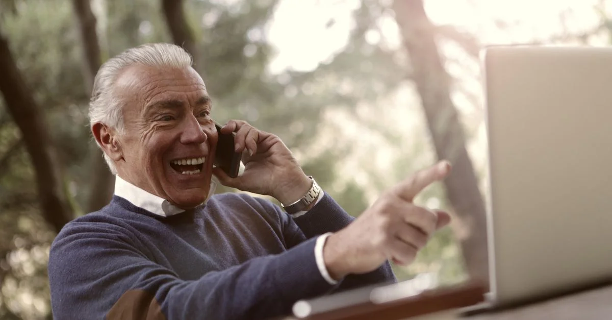 Un homme senior apprécie un appel téléphonique et utilise un ordinateur portable dans un cadre extérieur ensoleillé.