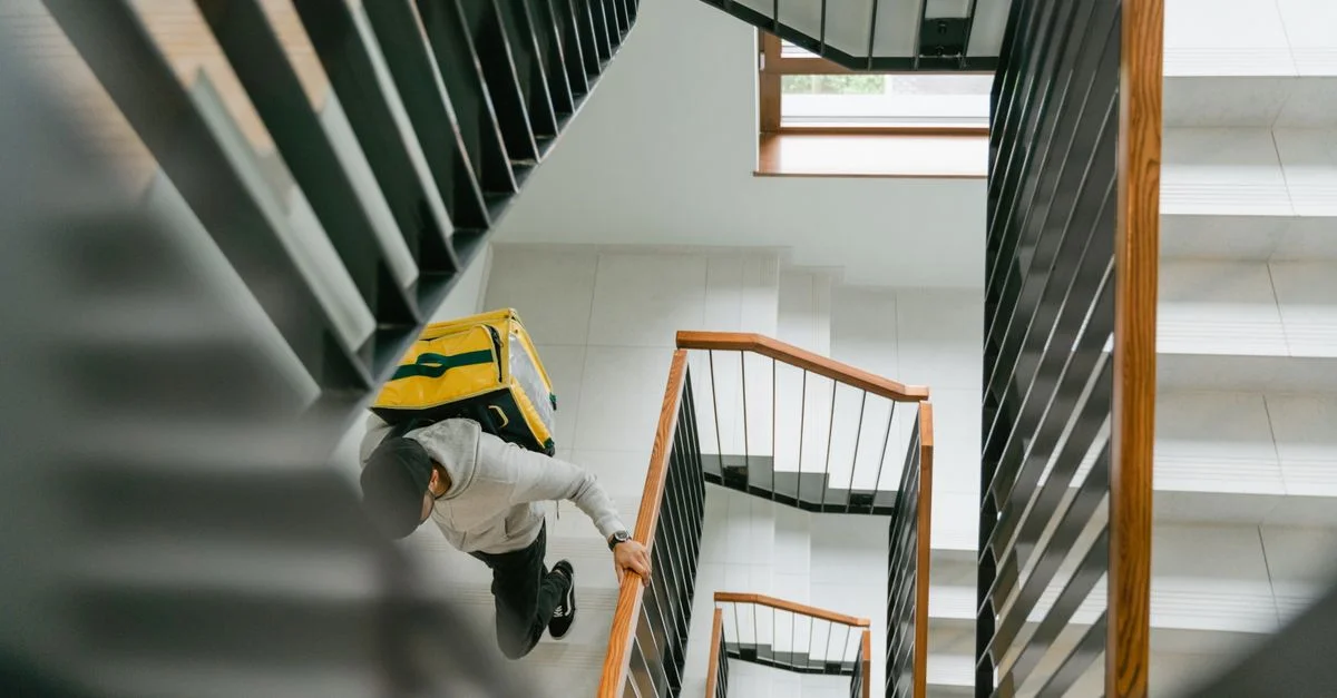 Un livreur portant un sac thermique jaune monte un escalier moderne à l'intérieur.