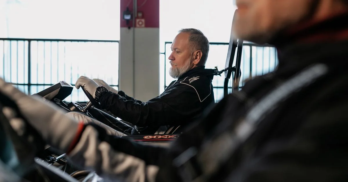 Un homme adulte senior en combinaison de course conduit un go-kart sur une piste intérieure, concentré sur la course.