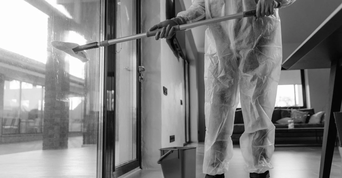 Une personne en combinaison de protection nettoie des portes en verre à l'intérieur, mettant l'accent sur l'hygiène.