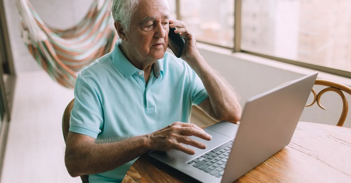 Homme âgé multitâchant avec un ordinateur portable et un téléphone à l'intérieur, démontrant l'utilisation de la technologie.