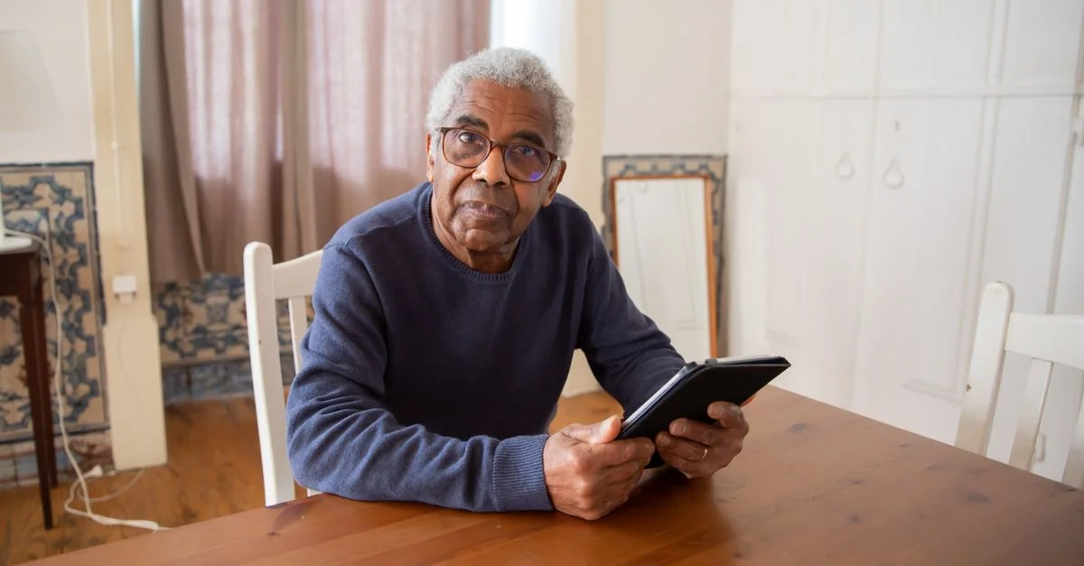 Homme africain-américain senior utilisant une tablette à la maison, assis à une table en bois, à l'intérieur.