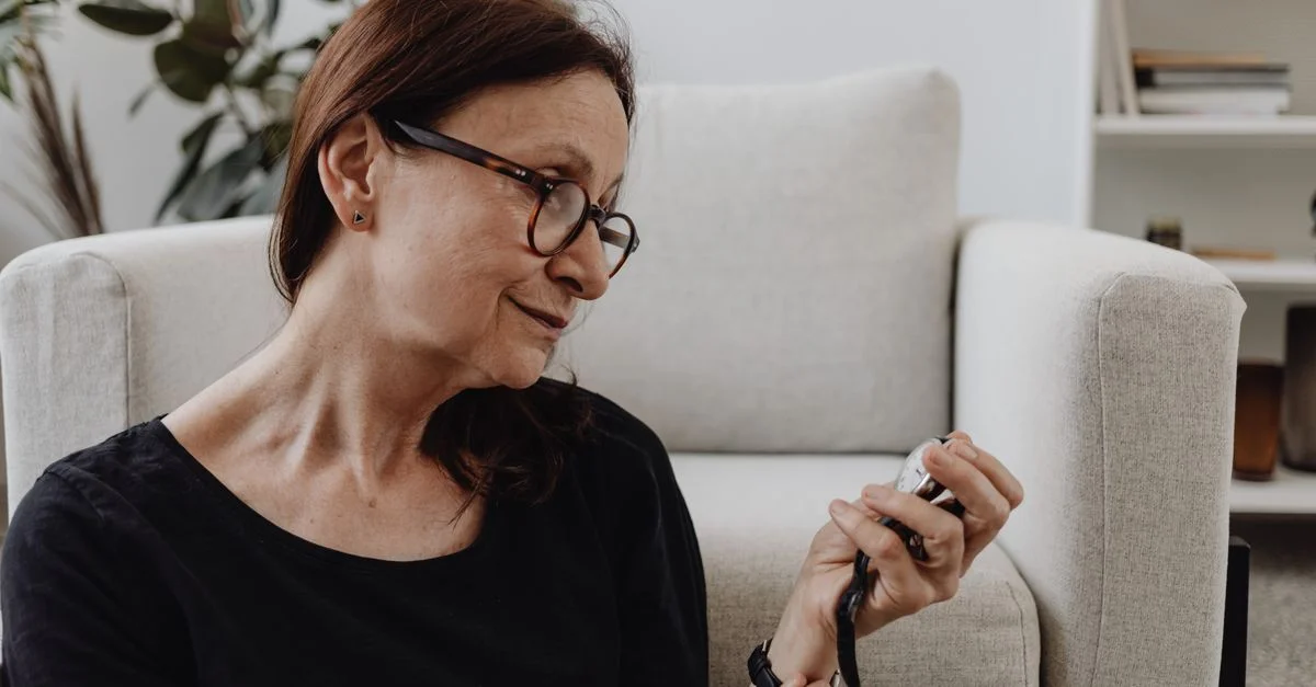 Femme senior réfléchie portant des lunettes et tenant une montre, en intérieur.