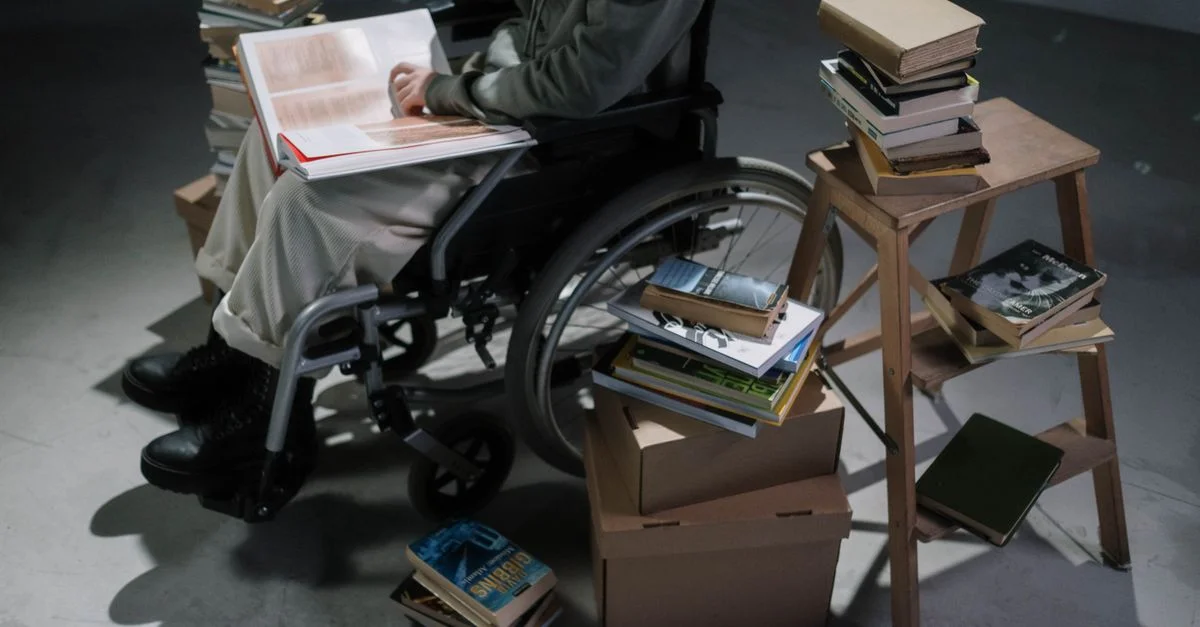 Une personne en fauteuil roulant lit un livre entourée de piles de livres à l'intérieur.