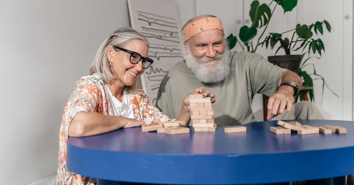 Un couple âgé jouant au Jenga à l'intérieur, profitant de moments de loisirs ensemble autour d'une table bleue.
