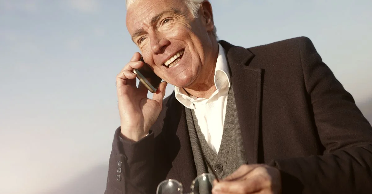 Homme senior en costume, souriant tout en parlant sur un smartphone, profitant de son temps libre à l'extérieur.