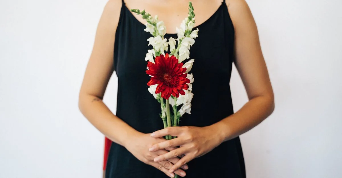 Une personne en robe noire tenant un bouquet de fleurs rouges et blanches sur un fond blanc épuré.