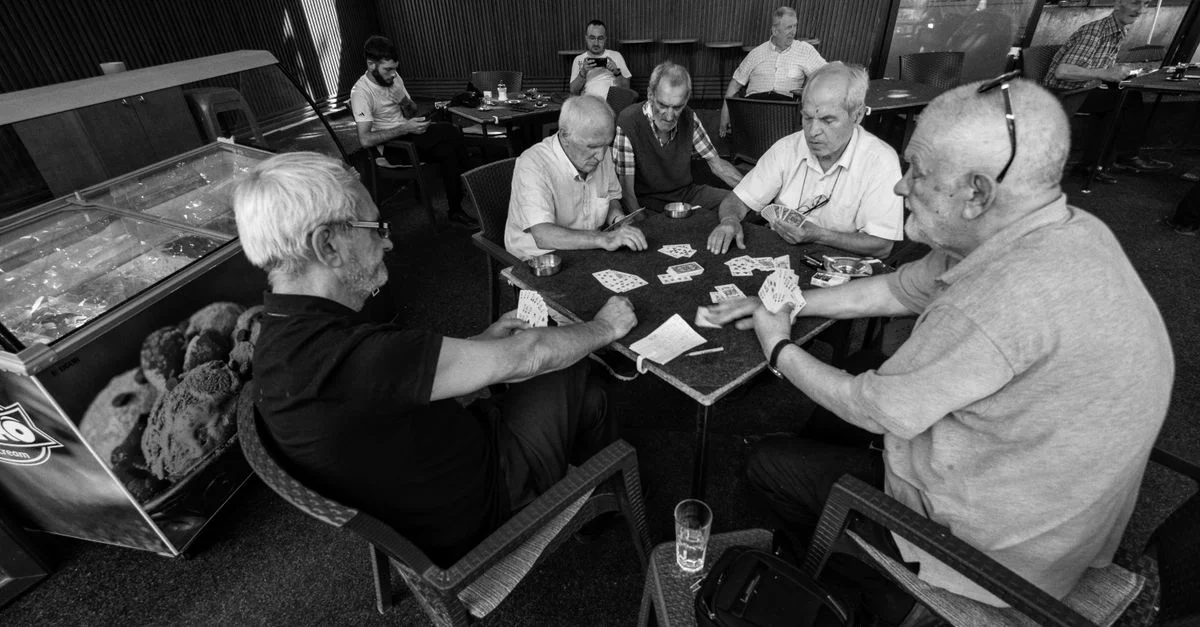 Photo en noir et blanc d'hommes âgés jouant aux cartes dans un café à Üsküp, créant une atmosphère détendue et sociale.