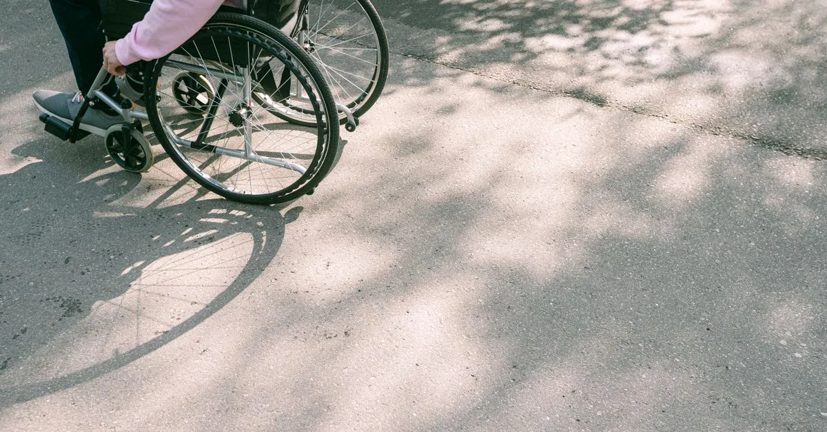Une personne en fauteuil roulant naviguant sur une route ensoleillée avec des ombres d'arbres. L'image capture un moment de mobilité et d'indépendance.